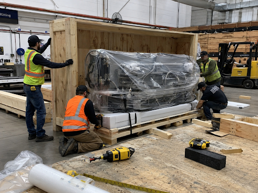 Workers in safety vests secure and pack large industrial machinery inside a custom wooden shipping crate in a warehouse.
