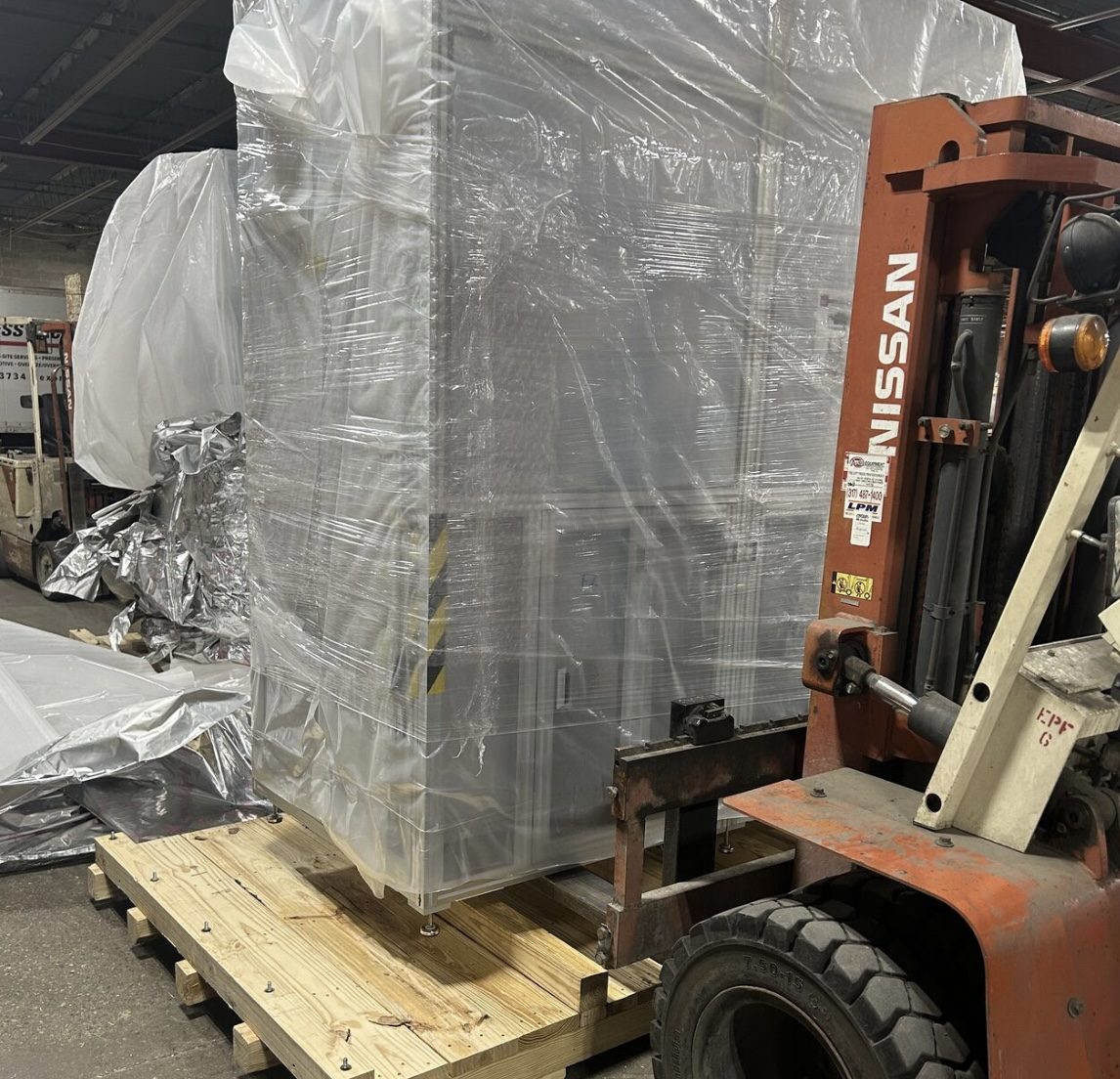 Large industrial machine wrapped in clear protective plastic on a wooden pallet beside a forklift inside a warehouse.