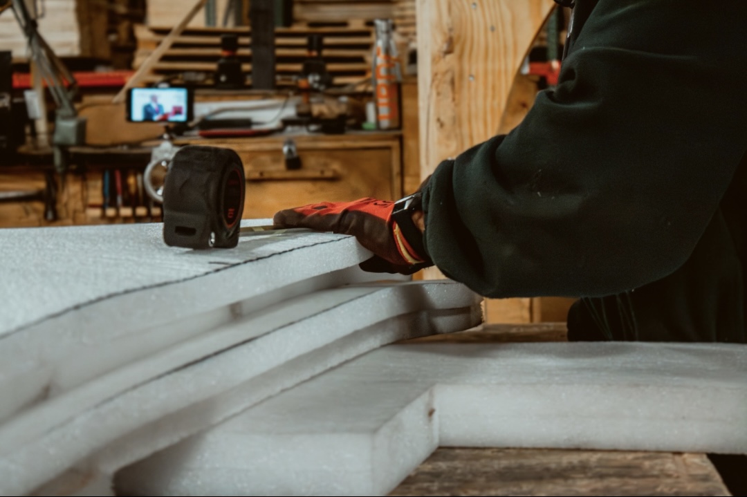 Worker wearing protective gloves measuring and preparing thick foam insulation boards on a workbench inside a workshop.